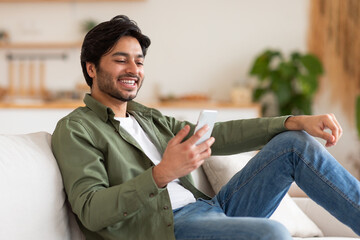 Wall Mural - A man is sitting on a couch in a home setting, smiling while looking at his smartphone. He is wearing a green shirt, white t-shirt, and blue jeans.