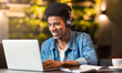 © Prostock-studio - A young black guy sits at a table in a coffee shop, wearing headphones and focusing intently on his laptop screen. He appears engrossed in his work or entertainment