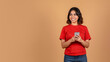 © Prostock-studio - A young middle eastern woman is standing against a plain beige background. She is wearing a bright red t-shirt and blue jeans. She is holding a smartphone in her hands and looking at copy space
