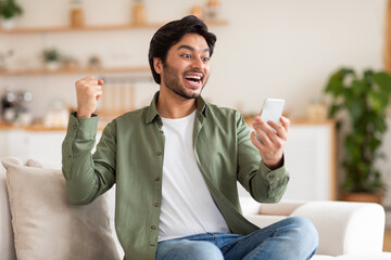 Wall Mural - A young man, dressed in a green shirt and jeans, sits on a white couch in his living room. He holding his smartphone in his right hand and celebrating, with his left fist in the air