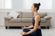 © Prostock-studio - A young woman sits in a meditative pose in her living room, her eyes closed and hands in a mudra.