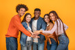 © Prostock-studio - This image shows a diverse group of five friends stacking their hands together in a studio setting. They are all smiling and appear to be enjoying each others company. The background is bright yellow