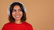 © Prostock-studio - A young middle eastern woman with dark brown hair smiles at the camera while wearing white headphones and a red t-shirt. The background is a light tan, copy space