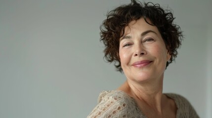 Wall Mural - A woman with short curly hair smiling gently wearing a light-colored lace top against a neutral background.