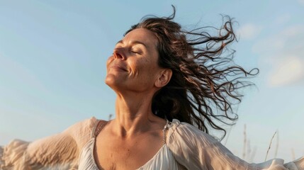 Wall Mural - Woman with closed eyes smiling wind blowing her hair standing in a field wearing a white blouse looking up to the sky.