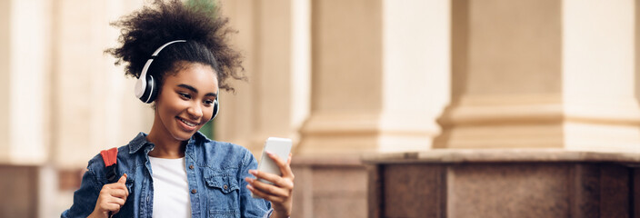 Wall Mural - Cheerful Black Student Girl Using Cellphone Texting And Browsing Internet, Wearing Headphones Standing Outdoor Near University Building. Listening To Educational Podcast Via Phone, Copy Space