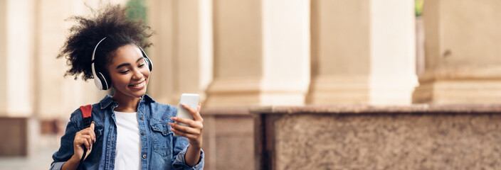 Wall Mural - Educational Podcast. African American Student Girl Using Phone Wearing Wireless Headphones Walking At University Campus Outdoor. People And Gadgets, E-Learning Application Concept, Copy Space