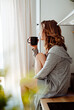 © ASTA Concept - Young pretty woman drinks coffee sitting at her kitchen at home and looking out the window enjoys of morning.