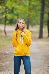 Wall Mural - Cheerful blue-eyed blonde girl in yellow hoodie, smiles against park background. Attractive blonde in stylish clothes posing outdoor.