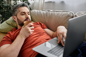 A bearded man in casual attire works on his laptop while reclining on a couch. He is holding a mug in one hand and appears to be focused on his work.
