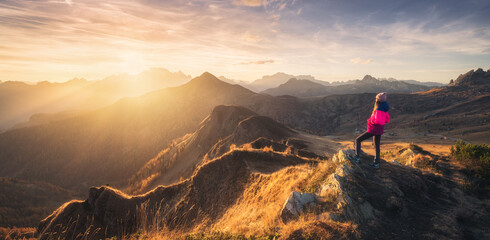 Wall Mural - Young woman on mountain peak and beautiful mountain valley in haze at colorful sunset in autumn. Dolomites, Italy. Sporty girl, mountain ridges in fog, orange grass, trees, golden sun in fall. Hiking