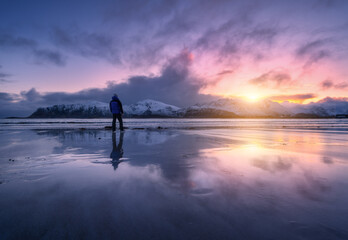 Wall Mural - man on the sandy beach against snowy mountains and colorful sky at sunset in winter. lofoten islands