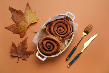 Wall Mural - Baking dish of tasty cinnamon rolls and autumn leaves on brown background