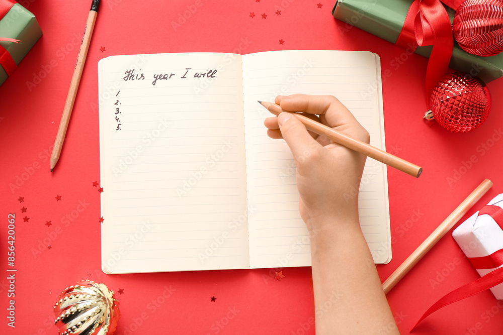 Woman writing THIS YEAR I WILL in notebook and Christmas decor on red background