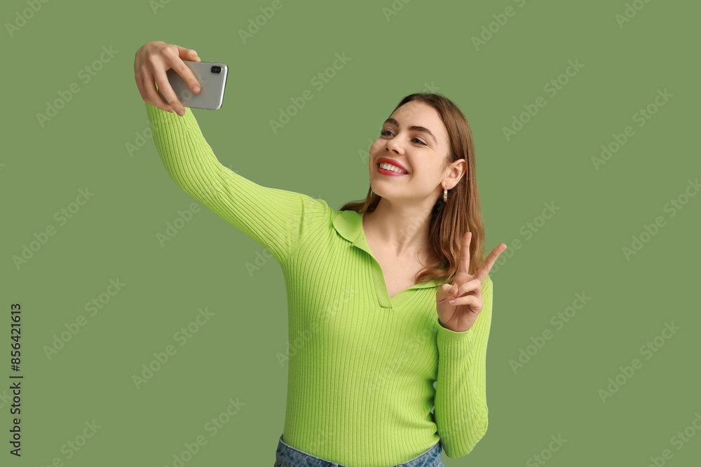 Pretty young woman showing peace gesture while taking selfie on green background