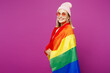 © ViDi Studio - Side profile view young happy European lesbian woman wrapped in striped rainbow flag looking camera isolated on plain purple wall background studio portrait. Pride day June month love LGBTQ concept.