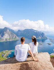 Wall Mural - A couple sits on a rocky cliff overlooking a stunning Norwegian fjord landscape. The azure waters, lush green mountains, and clear blue sky create a picturesque backdrop. Reinebringen, Lofoten, Norway