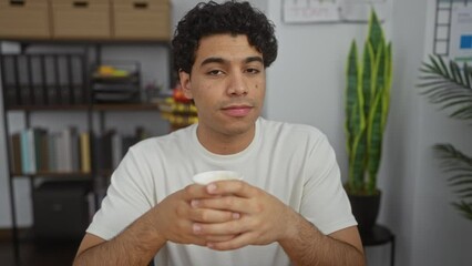 Canvas Print - Young hispanic man seated in an office, holding a cup while looking contemplative in a modern indoor workplace with plants and shelves in the background.