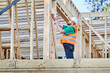 © anatoliy_gleb - Carpenter constructing two-story wooden frame house near the forest. Bearded man in glasses hammering nails into structure while wearing protective helmet and construction vest.