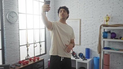 Sticker - Young man making a video call while flexing his arm in a gym, surrounded by various workout equipment in an indoor sports center.