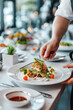 © Irina B - Hand arranging a gourmet dish with various vegetables and microgreens on a white plate in a contemporary restaurant..