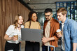 © LIGHTFIELD STUDIOS - Multicultural colleagues in casual attire gather around a laptop for a collaborative tech discussion during a corporate trip.