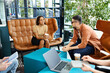 © LIGHTFIELD STUDIOS - Multicultural colleagues in casual clothes work together around a table in a hotel lobby during a corporate trip.