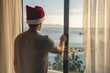 © alisaaa - Man in hotel room celebrating chritmas, he opens the curtains in the morning looking at the sea view, he is wearing a santa hat Man in hotel room celebrating chritmas, he opens the curtains in the mor