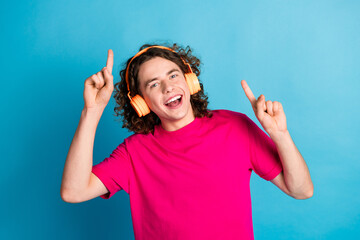 Poster - Photo of funky guy with wavy hair earrings dressed pink shirt raising fingers listen music in headphones isolated on blue color background