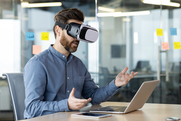 Wall Mural - A young businessman works in the office, sits at a table with a laptop, wears a virtual mask and communicates online via video communication, gesturing with his hands