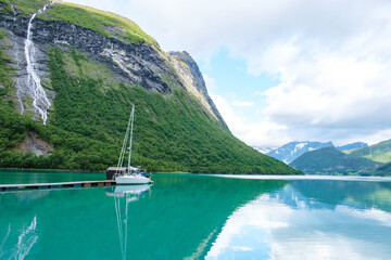 Wall Mural - A white sailboat is docked at a wooden pier, nestled in a picturesque Norwegian fjord surrounded by lush green mountains and a cascading waterfall. Norangsfjorden Urke Norangdal Norway
