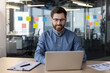 © Tetiana - A smiling young man in glasses and a blue shirt sits in the office at the desk and concentrates on using a laptop, typing on the keyboard