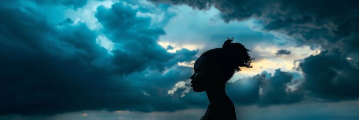 Wall Mural - A silhouette of a black woman stands against a backdrop of dramatic storm clouds, creating a striking contrast between light and dark