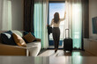 © shine - Confident businesswoman wearing white shirt and holding suit with suitcase stands in a hotel room while looking at the view from window Beautiful sunset. Woman enjoying her holiday in a luxury hotel.