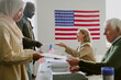 © AnnaStills - Side view shot of senior polling station workers giving ballot papers to ethnically diverse young voters