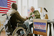 © AnnaStills - Senior Caucasian polling station worker giving ballot paper to young African American voter in wheelchair
