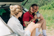 © Ilona - Two women friends are sitting in the back of car, talking and drinking coffee from a thermos during a trip out of city.