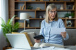 © Liubomir - Senior woman looking worried while managing finances at home, using a laptop and holding documents. Concept of financial stress, budgeting, and retirement planning.