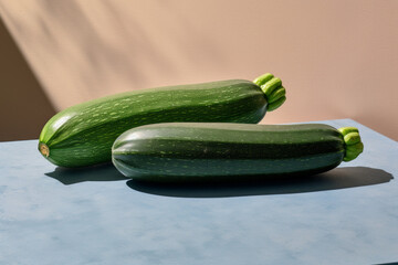 Two fresh zucchinis on a light blue and beige surface with shadows.