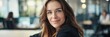 © Elmira - A mid-range shot of a Caucasian businesswoman sitting at a desk in a corporate office setting. She is smiling and looking directly at the camera with confidence