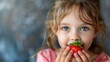 © Pinklife - A young girl with blue eyes and wavy hair enjoys eating a large red strawberry, capturing an innocent and joyful moment in her childhood, the contrast of red and blue enhancing the image.