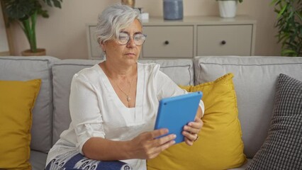 Canvas Print - Mature woman with grey hair in glasses sits on a sofa in living room, focused on using a blue tablet device, surrounded by stylish home decor with yellow pillows and indoor plants.