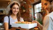 © Pinklife - A woman with a white headscarf and apron smiling as she serves pizza to a customer in a cozy restaurant setting, showcasing hospitality and freshness of the food.