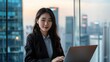 © buraratn - Asian businesswoman with laptop in a high-rise office - A successful Asian businesswoman working on a laptop, with a panoramic city view in the background, showcasing a high-rise office setting