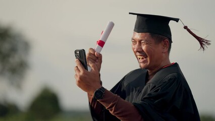 Wall Mural - A man in a graduation gown is holding a cell phone and laughing. He is taking a picture of himself with his graduation cap and gown