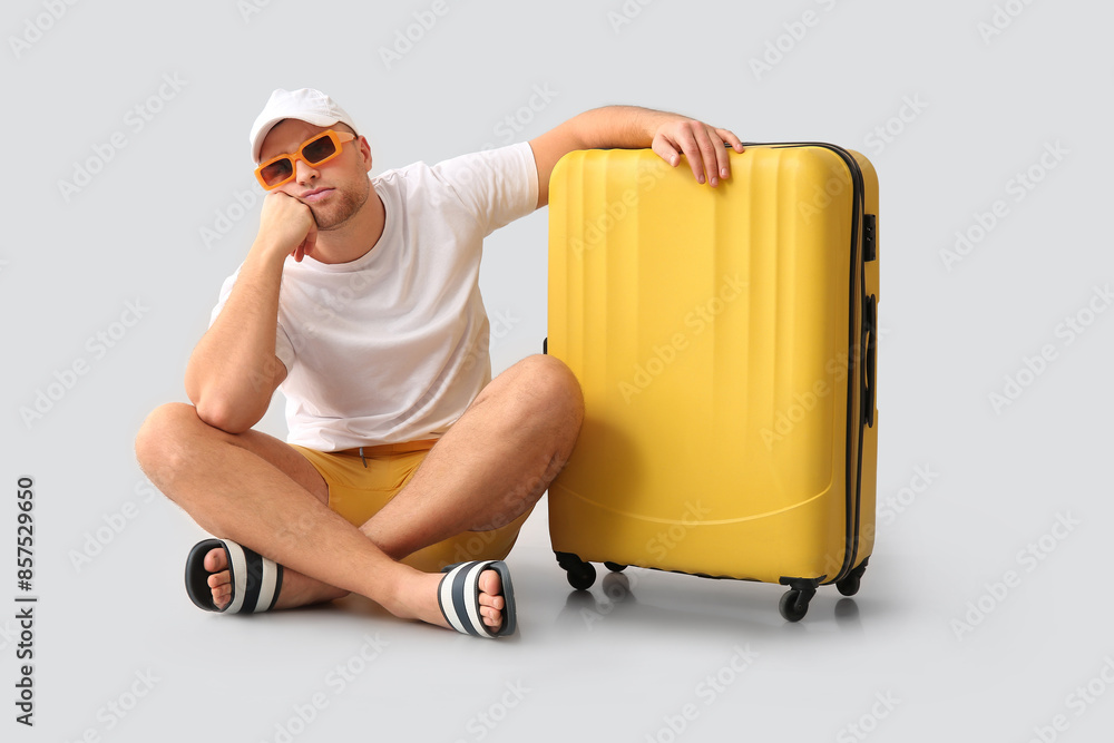 Handsome young bored male tourist with suitcase sitting on grey background
