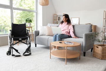 Poster - Young African-American woman with laptop and wheelchair sitting on sofa at home