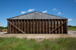 © Kolanderverse - Brown Storage Facility Shed - Simple Wood Structure for Storing Cement, Sand, Salt, Fertilizer, Seed, Feed, or Straw Against a Blue Sky