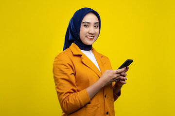Wall Mural - Happy young Asian woman holding mobile phone, looking at camera and smiling, texting or browsing internet isolated on yellow background