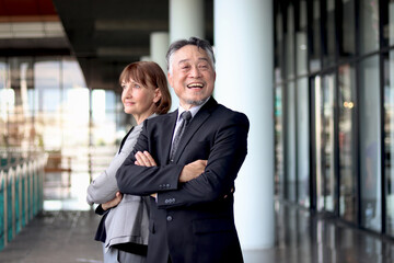 Happy smiling senior Asian businessman in suit with Caucasian businesswoman standing with arms crossed at office. Elderly successful male and female boss at workplace. Portrait  mature businesspeople.